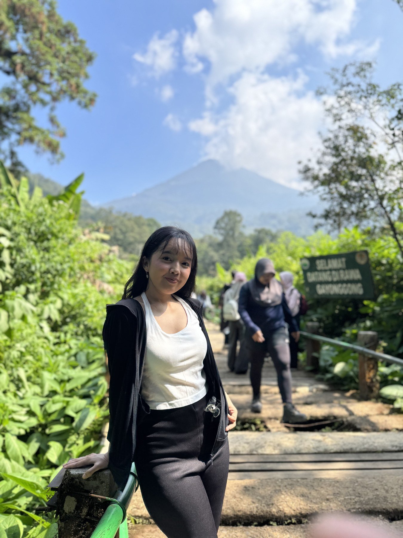 Dea berdiri di jalan taman dengan pegunungan dan langit biru di belakangnya